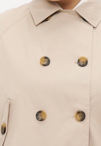 Beige dubbelknäppt trenchcoat tillverkad av slät tyg, med rundade bruna knappar med marmormönster och en strukturerad krage.