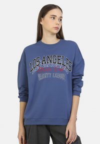 Sudadera azul con cuello redondo, corte holgado y texto impreso "LOS ÁNGELES Athletic Dept Varsity League" en blanco y rojo.