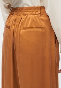 Καφέ φαρδιά παντελόνια με ελαστική μέση και πίσω τσέπες με σχισμές, φορεμένα με κρεμ μπλούζα μακρυμάνικη.