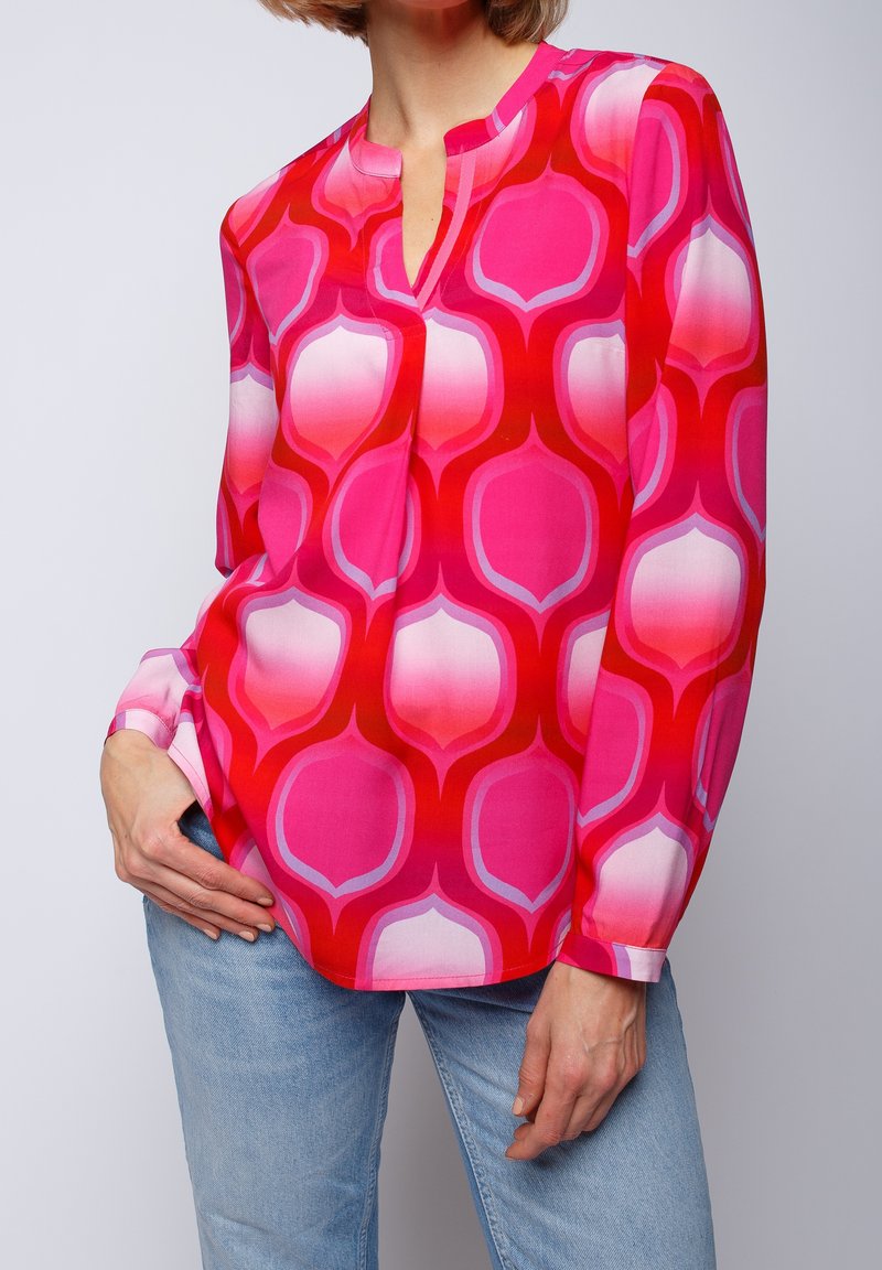 Langærmet bluse i stærk pink med et geometrisk mønster af dristige røde og lyserøde former. Har en v-udskæring og knapmanchetter.