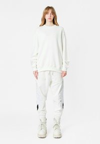 Sudadera blanca con cuello redondo, combinada con pantalones deportivos blancos que presentan patrones de alas plateadas. El calzado es voluminoso y con cordones, también en blanco.