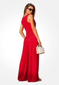 Mono rojo sin mangas con un diseño fluido y una cintura ajustada. La modelo sostiene un pequeño bolso beige y lleva gafas de sol.