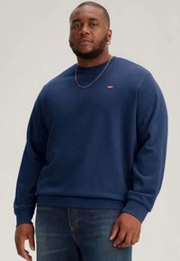 Navy blue sweatshirt made of cotton blend, featuring a crew neckline and ribbed cuffs. Small red logo on the chest, worn with blue jeans.