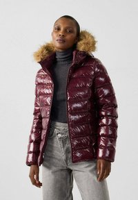 Burgunderfarbene Pufferjacke mit glänzender Oberfläche, ausgestattet mit einer mit Fell gefütterten Kapuze, Reißverschluss und horizontalem Steppmuster; getragen über einem dunklen Rollkragenpullover.