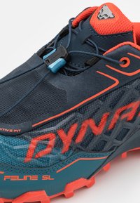 Chaussure de trail running bleu foncé avec des accents orange, tige en maille, système de laçage rapide et semelle extérieure texturée. Présente les marques "DYNAFIT" et "FELINE SL".