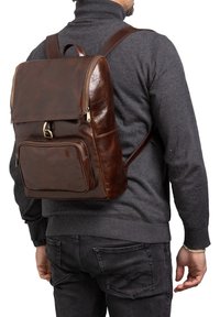 Zaino in pelle marrone con flap superiore con zip, tasca frontale e spallacci regolabili. Texture liscia con un leggero luccichio e cuciture.