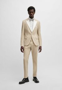 Beige Zweiteiler mit weißen Schalklapellen, schmal geschnittene Hose, kombiniert mit einer grauen Fliege und schwarzen Schuhen. Glatte Stofftextur.
