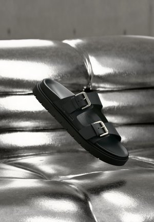Sort slidesandal med to justerbare spænder, svævende over en skinnende metallisk sølv tuftet overflade.