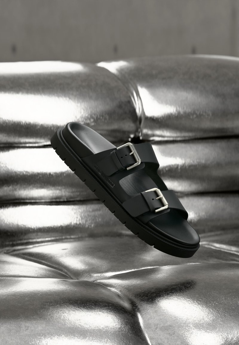 Μαύρο σανδάλι slide με δύο ρυθμιζόμενες αγκράφες που αιωρείται πάνω από μια γυαλιστερή μεταλλική ασημένια επιφανειακή επιφάνεια με ραφές.