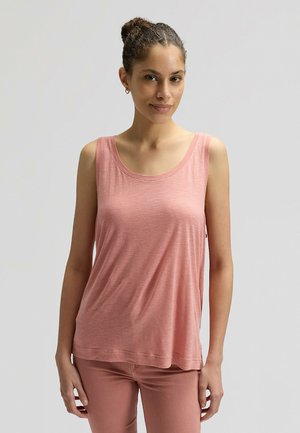 Giovane donna con capelli ricci raccolti in uno chignon, che indossa un top senza maniche rosa e pantaloni rosa, in piedi davanti a uno sfondo chiaro semplice.