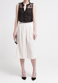 Blouse noire sans manches avec superposition en dentelle et détails de boutons, associée à un pantalon blanc à jambes larges. Escarpins noirs à bout pointu et pochette.