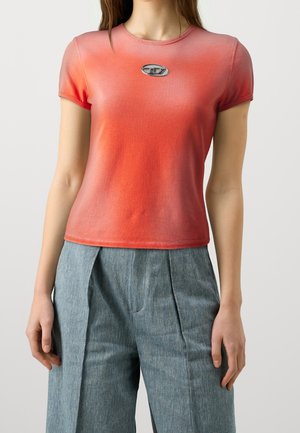 Frau trägt kurzärmeliges, verblasstes rot-orangefarbenes T-Shirt mit Metalllogo auf der Brust und hoch taillierte blau-graue plissierte Hose.