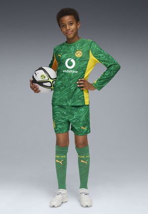 Zielony strój piłkarski z żółtymi akcentami, posiadający teksturowany wzór, logo i dopasowane spodenki. Zawodnik trzyma czarno-białą piłkę nożną.