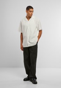 Camicia bianca a maniche corte con un motivo a strisce orizzontali testurizzate, abbinata a pantaloni a fantasia scura e scarpe nere.