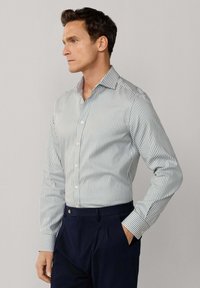 Mann trägt ein hellblau-weiß gestreiftes Hemd mit langen Ärmeln, das in dunkelblaue, plissierte Hosen gesteckt ist, eine Hand in der Tasche.