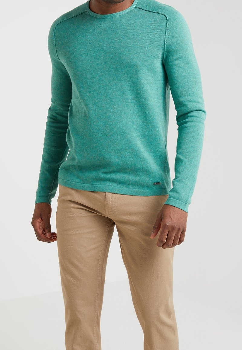 Turkos långärmad tröja med rund halsringning, tillverkad av ett texturerat material, matchad med ljusbeige byxor. Har sömnadsdetaljer på axlarna.