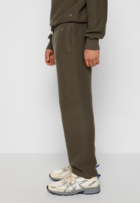 Olivgrüne, strukturierte Jogginghosen mit geripptem Bund, seitlichen Taschen und subtilen Details; kombiniert mit beigen und grauen Sportschuhen.