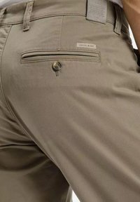 Beżowe spodnie o gładkiej fakturze, z tylną kieszenią zapinaną na guzik i metką z napisem "CROSS JEANS". Widoczne są subtelne detale przeszycia.