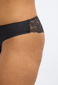 Μαύρο στρινγκ με διακόσμηση δαντέλας και λείο ύφασμα στο σώμα. Η μέση διαθέτει περίπλοκη λεπτομέρεια δαντέλας, προσφέροντας μια απαλό αντίθεση με το κύριο υλικό.