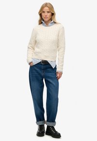 Crème kabelgebreide trui met geribde manchetten, gecombineerd met denim jeans en zwarte veterschoenen. Onder de trui is een blauw gestreept overhemd met kraag zichtbaar.