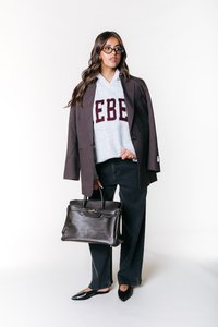 Szara bluza z kapturem z czerwonymi literami, brązowa marynarka w kratę, czarne spodnie typu wide-leg i czarne baleriny. Trzyma ciemną skórzaną torebkę z złotymi akcentami.