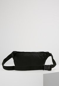 Svart tyg midjeväska med justerbar rem och sidofästspänne, placerad på vit yta mot en enkel ljusgrå bakgrund.