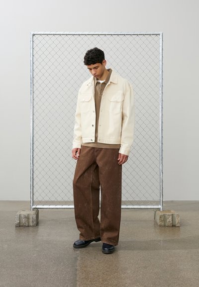 Bărbat tânăr stând în interior, purtând o geacă bej, o cămașă maro, pantaloni largi maro și pantofi negri, în fața unui cadru metalic cu lanțuri.