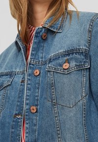 Jeansjacka i ljusblått med två bröstfickor, kopparfärgade knappar och en klassisk krage. Tyget har en strukturerad yta.