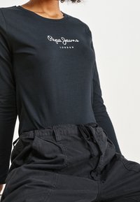 Černé tričko s dlouhým rukávem a kulatým výstřihem, na hrudi s bílým logem "Pepe Jeans London", v kombinaci s volnými černými kalhotami.