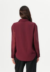 Femme aux longs cheveux foncés portant un chemisier bordeaux transparent à manches longues et un pantalon noir, vue de dos sur un fond uni.