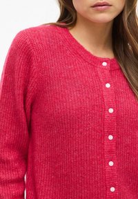 Roza pletena jakna s krožnim izrezom, sedmimi belimi gumbi spredaj in teksturirano, mehko tkanino z rahlim sijajem.