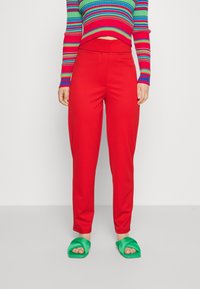 Femme portant un pantalon rouge taille haute, un pull cropped à rayures rouges, bleues et vertes, et des sandales vertes sur un sol gris.