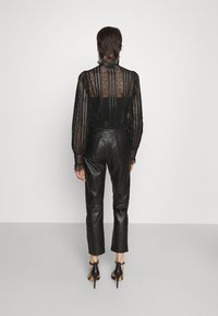 Blouse en dentelle noire à manches longues avec des motifs détaillés ; associée à un pantalon en cuir noir ajusté et des chaussures noires à talons hauts.