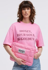Pink gepolsterte Laptophülle mit gesteppter Textur, versehen mit einem vorderen Etikett, auf dem "SENSES .THE LABEL." steht. Das Modell trägt ein pinkfarbenes T-Shirt mit Grafikaufdruck.