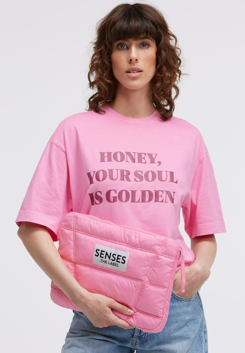 Pink gepolsterte Laptophülle mit gesteppter Textur, versehen mit einem vorderen Etikett, auf dem "SENSES .THE LABEL." steht. Das Modell trägt ein pinkfarbenes T-Shirt mit Grafikaufdruck.