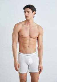 Bijele pamučne bokserice s uskim krojem, s mekanim elastičnim pojasom označenim "Calvin Klein." Glatka tekstura, minimalni dizajn.