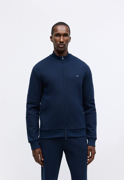 Modra zip-up jakna z visokim ovratnikom, rebrasto teksturo, dolgimi rokavi in logotipom na prsih; v kompletu z usklajenimi hlačami.