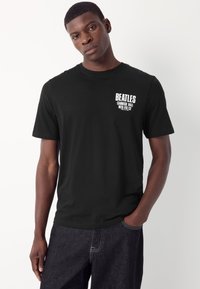 Black cotton t-shirt featuring a graphic print on the left chest declaring "BEATLES CARNEGIE HALL WED. FEB. 12" in white text. Short sleeves.