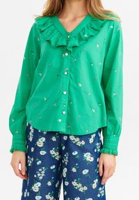 Camicia verde con colletto arricciato, ricami floreali e maniche lunghe. Abbinata a pantaloni blu con motivo floreale.