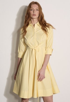 Femme aux longs cheveux roux portant une robe chemise à rayures jaunes et blanches avec des manches 3/4 et une ceinture nouée à la taille, debout devant un fond uni.