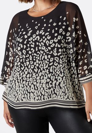 Kvinde iført en sort gennemsigtig bluse med hvidt abstrakt leopardmønster og stribet kant, kombineret med sorte bukser med læderlook.