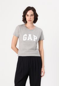 T-shirt gris à manches courtes avec col rond et logo "GAP" blanc. Le t-shirt est en tissu doux, associé à un pantalon plissé noir.