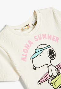 Valkoinen ribbattu lyhythihainen T-paita, jossa on vaaleanpunaisella painettuna "ALOHA SUMMER" ja piirretty koira sinisessä lippiksessä pitelemässä vaaleanpunaista surffilautaa.