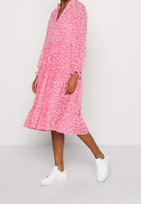 Abito floreale rosa con frontale a bottoni, maniche lunghe e orlo arricciato. Abbinato a sneakers bianche, su uno sfondo semplice.