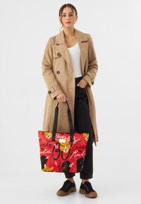 Rode totebag met grafische print met zwarte en gele patronen. Zwarte handvatten en een ritssluiting. Draagt bij een beige trenchcoat.