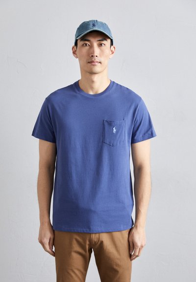 Tricou din bumbac albastru, cu un buzunar și un logo brodat pe piept, asortat cu pantaloni maro și o șapcă bleu deschis.
