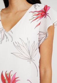 Blusa in tessuto leggero con motivo floreale, con scollo a V e maniche corte a palloncino. Presenta fiori rosa e grigi su uno sfondo bianco.
