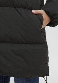 Zwarte pufferjack met een gladde, matte textuur. Beschikt over een zijzak, genaaide quiltontwerp en een verstelbare zoomkoord.