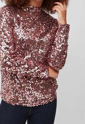 Femme portant un haut à manches longues et col montant recouvert de sequins brillants roses et or rose, avec une main levée près du visage.