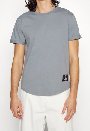 Man wearing a plain gray short-sleeve t-shirt with a small black Calvin Klein logo patch near the hem, paired with white pants.